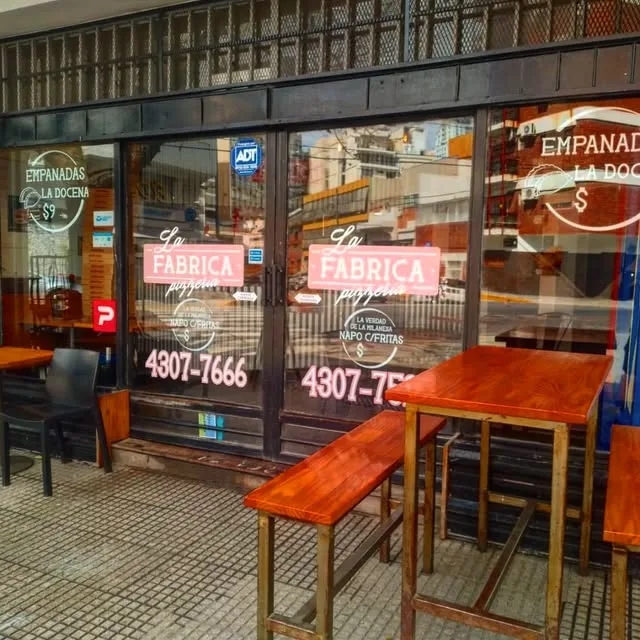 Frente de Pizzería La Fábrica en Azara 100, con el cartel en las vidrieras y mesas altas en la vereda